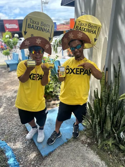 Dupla da OxePay segurando as placas Taxa Bem BAIXINHA